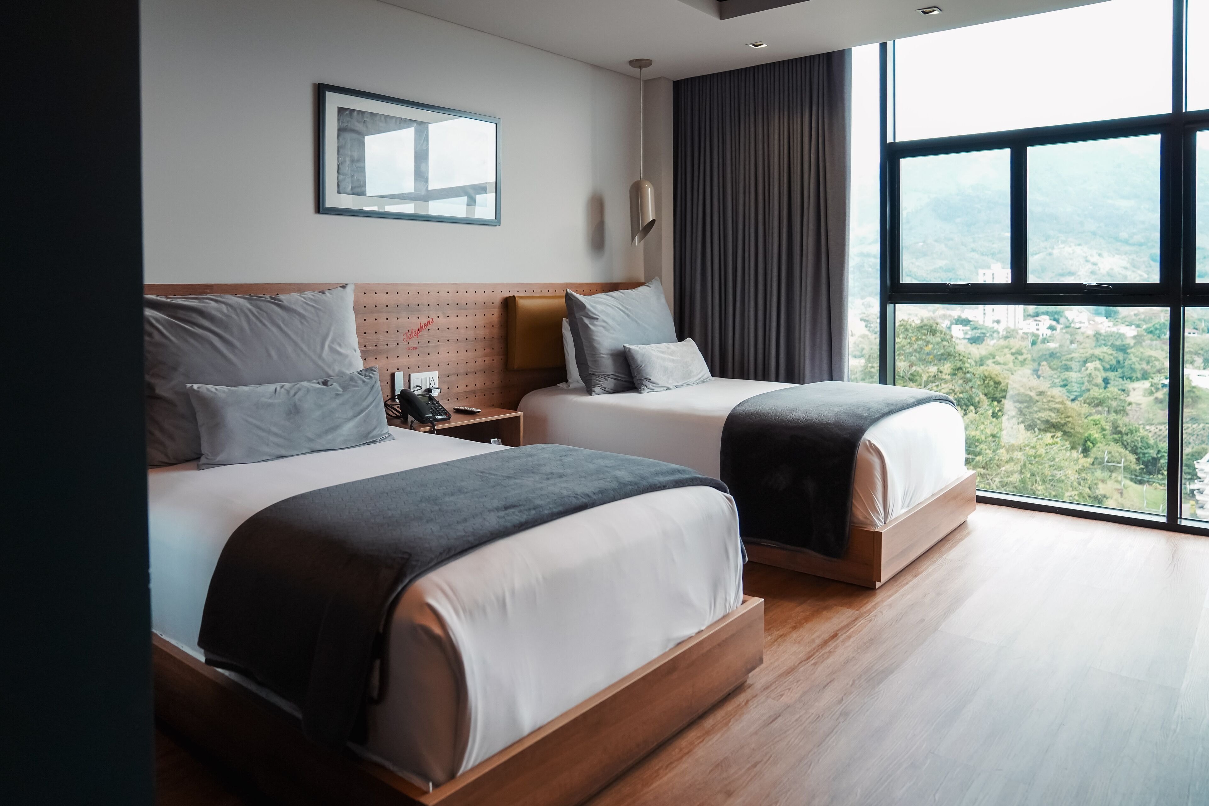 Une chambre d’hôtel moderne avec deux lits, une grande fenêtre donnant sur la ville, une œuvre d’art accrochée au mur et une table de chevet avec un téléphone.