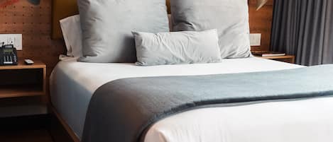 A modern hotel room with a wooden headboard, a large bed with grey bedding, a framed artwork on the wall, and a bedside telephone.