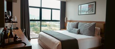 A modern hotel room with a large bed, a desk with wine bottles, and a view of the outdoors.