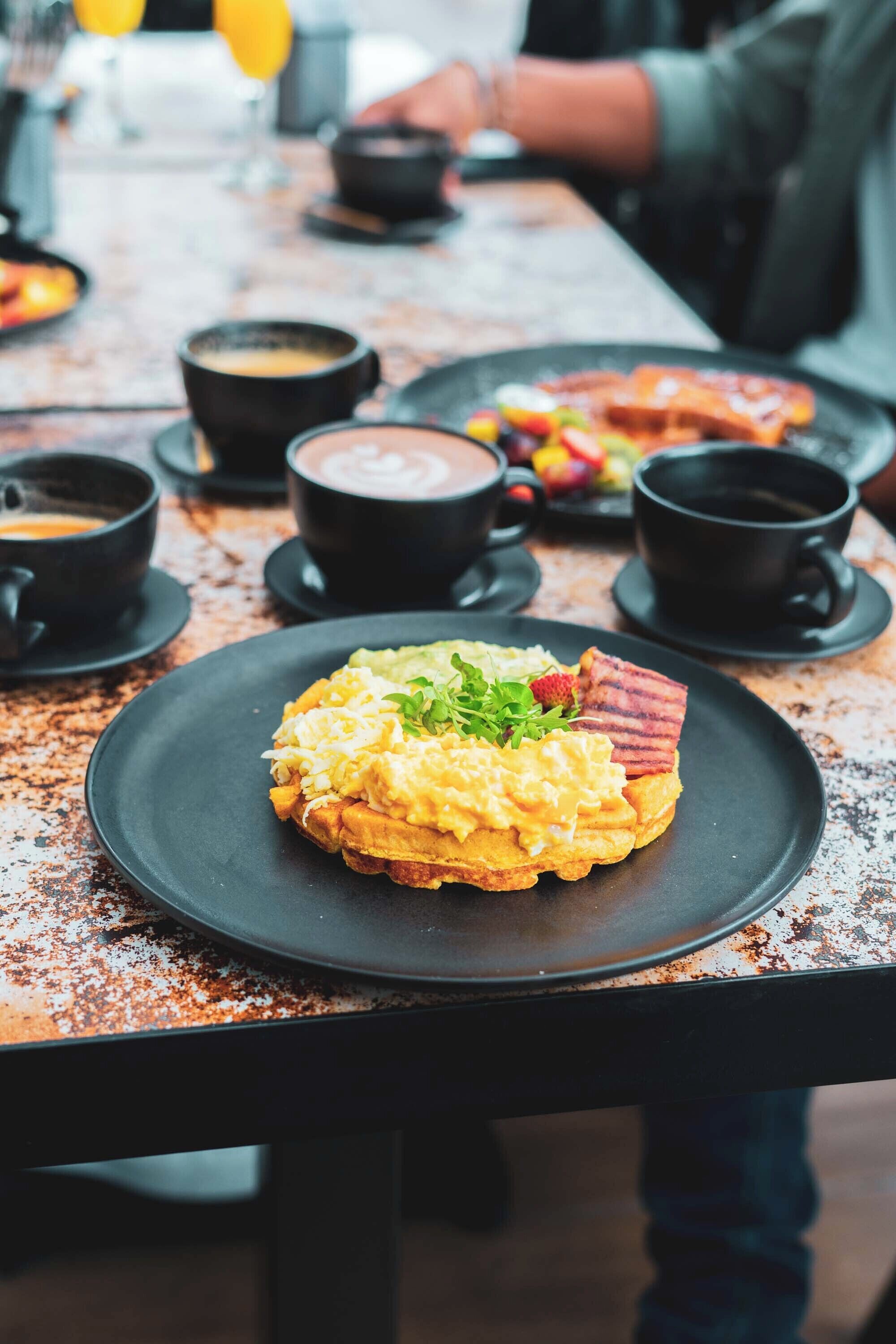 A breakfast setting with a plate of scrambled eggs, bacon, and greens on a black plate.