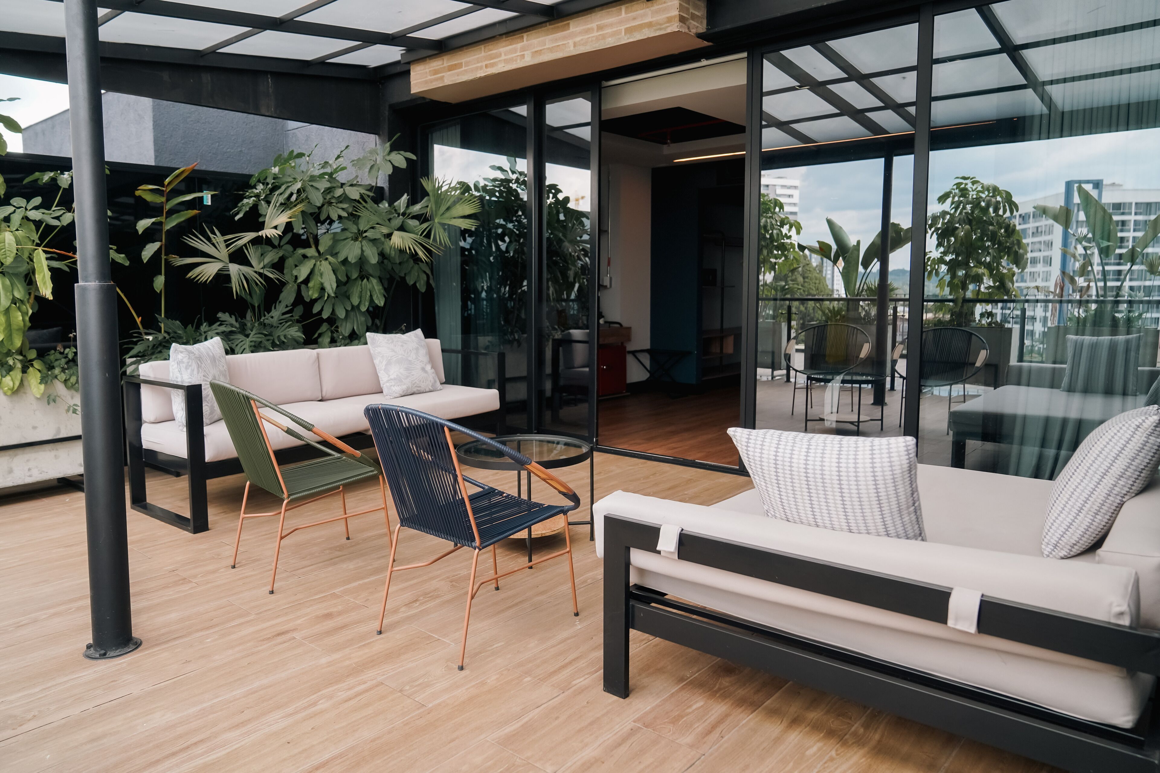 A modern outdoor seating area with a sofa, chairs, and a glass table, surrounded by large windows and indoor plants.