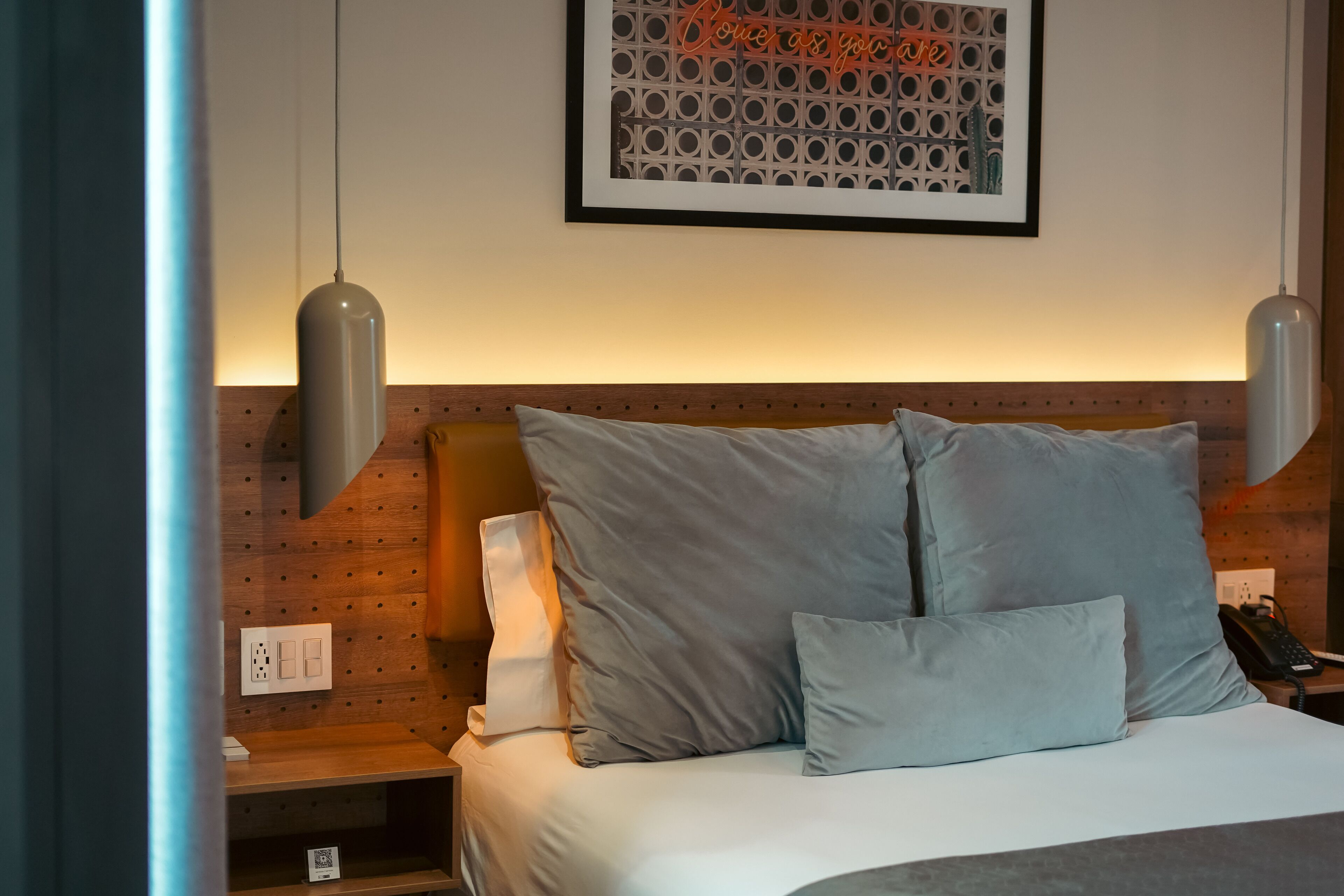 Een net opgemaakt bed met een hoofdbord, een bedlampje en een kunstwerk in een lijst aan de muur.