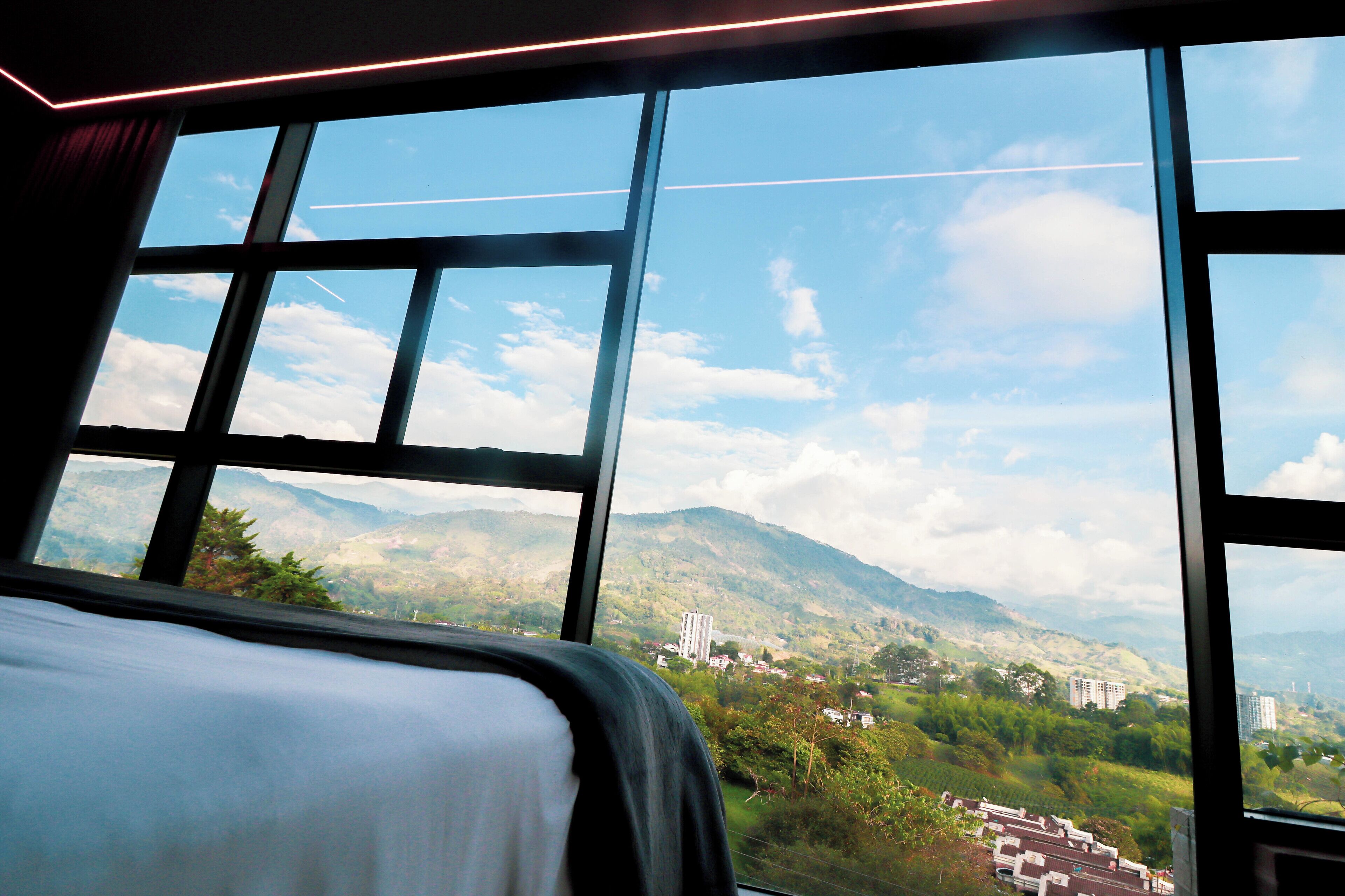 Habitación de hotel con un amplio ventanal que ofrece vistas a un paisaje montañoso y edificios al lejano.