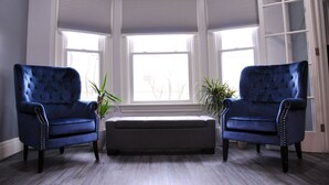 Une pièce avec deux fauteuils bleus, un banc et une grande fenêtre avec des volets.