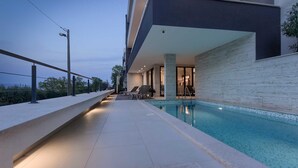 Zona moderna de piscina exterior con vistas al mar, un área de estar cubierta y un camino bien iluminado.