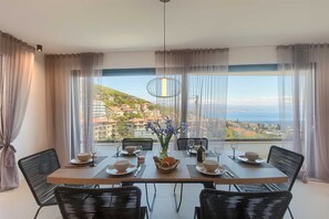 Uma área de refeições com uma mesa posta para quatro pessoas, um vaso de flores e vista para uma cidade costeira.