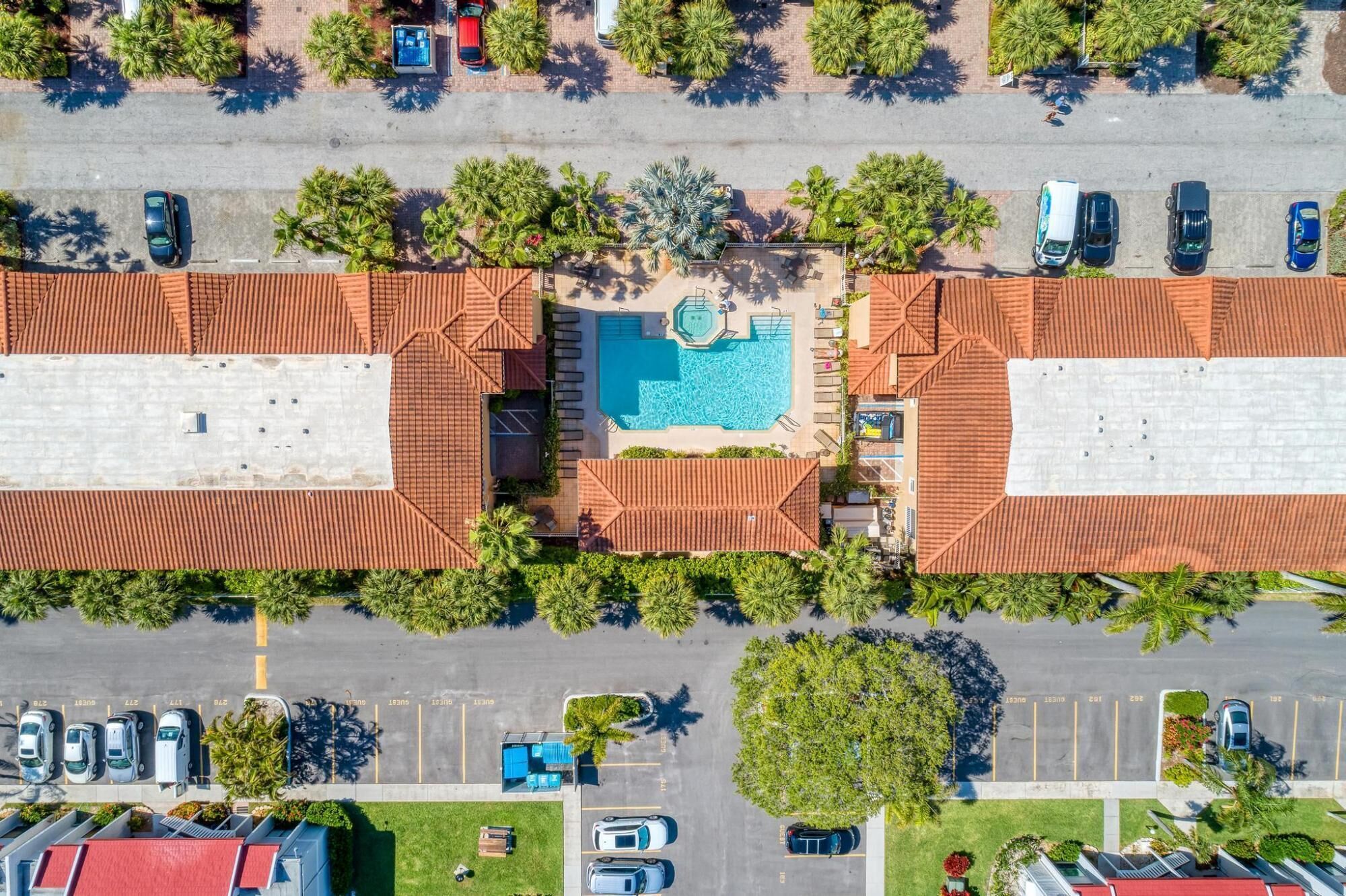 An aerial view of a hotel with a central swimming pool, surrounded by palm trees and a parking lot.