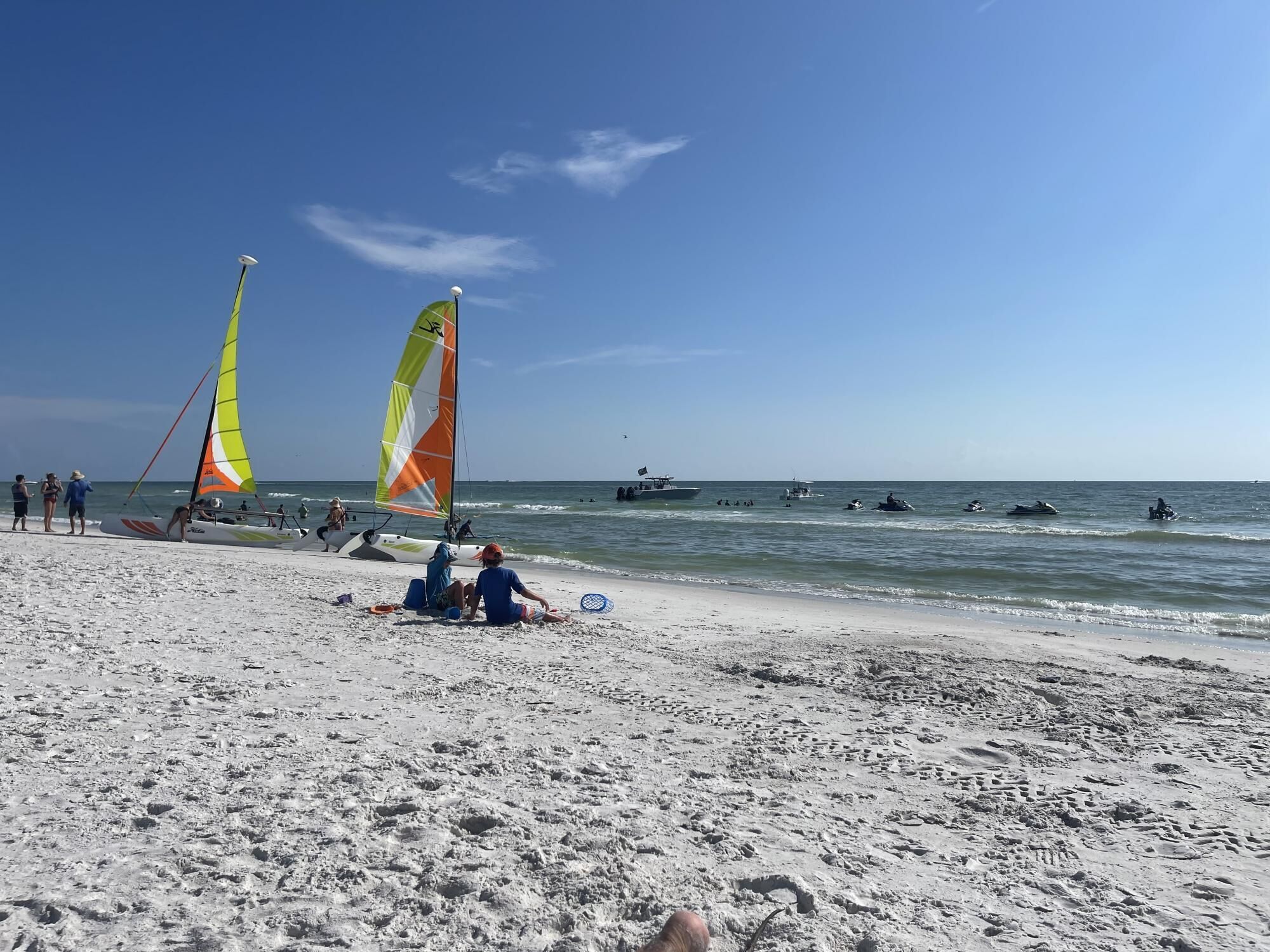 A beach with two sailboats, people relaxing on the sand, and several boats in the water.
