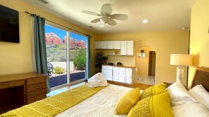 Habitación de hotel con cama, escritorio, silla, ventana con vista a formaciones rocosas rojas y ventilador de techo.