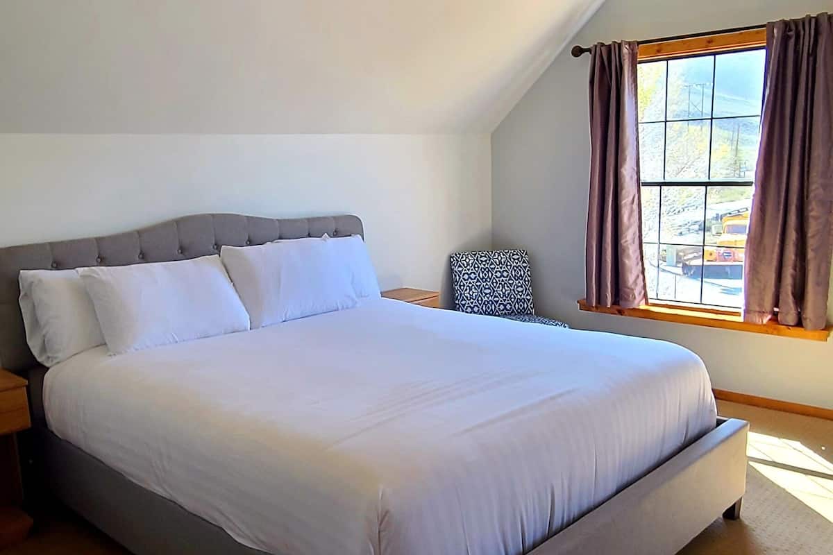 Uma cama grande com estrutura cinza e roupa de cama branca num quarto com teto inclinado e janela com cortinas.