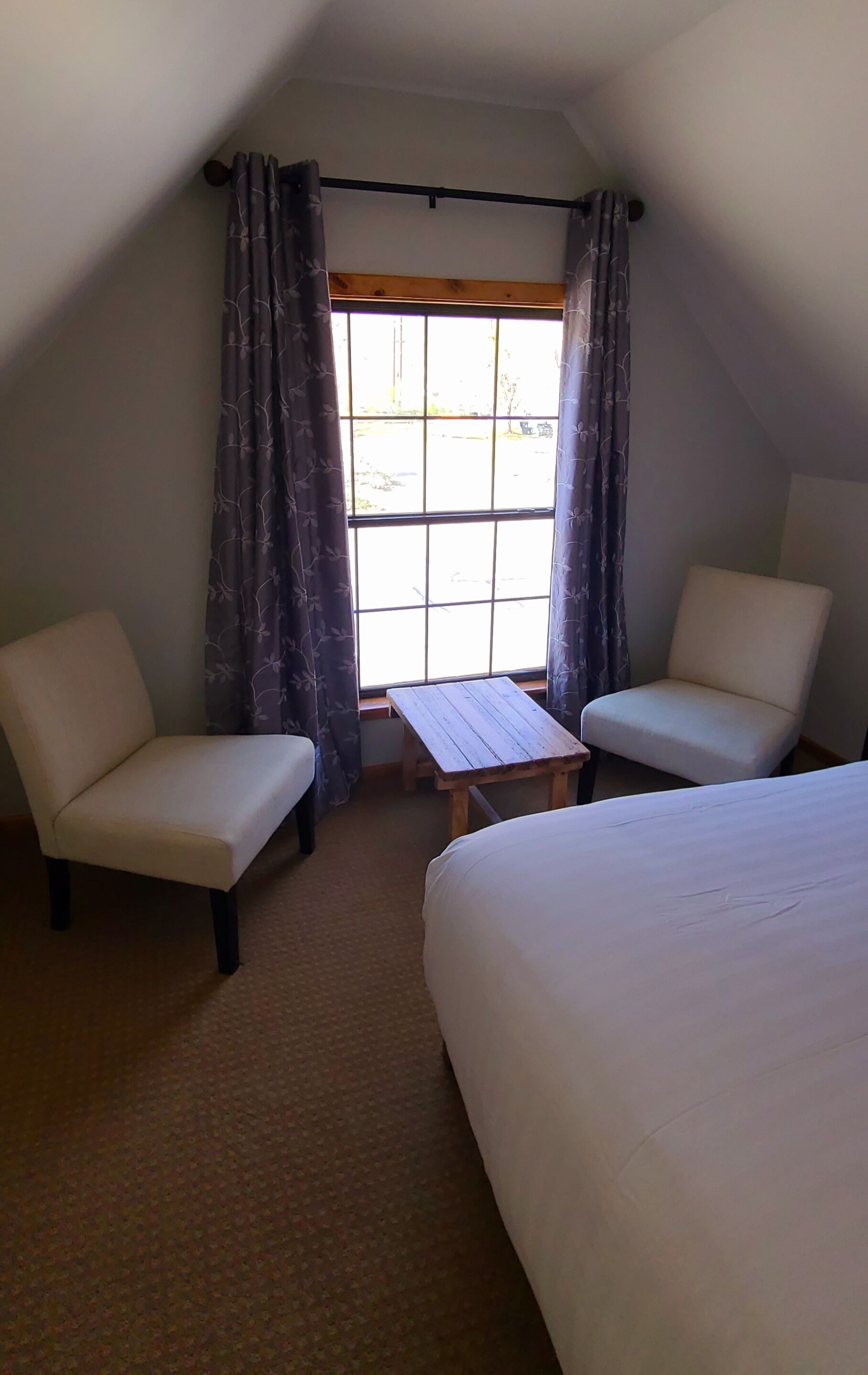 A room with two white beds, a small wooden table, two white chairs, and a window with curtains.