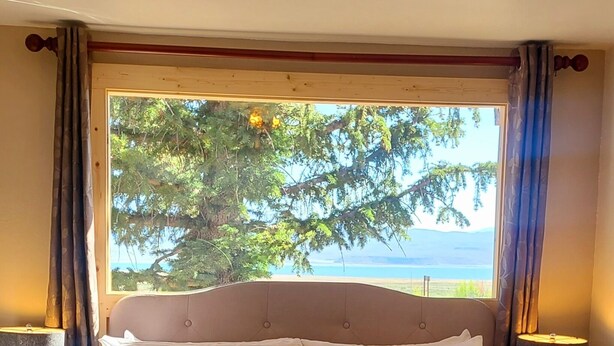 A bedroom with a large bed, wooden headboard, bedside lamps, and a view of trees and water through a window.