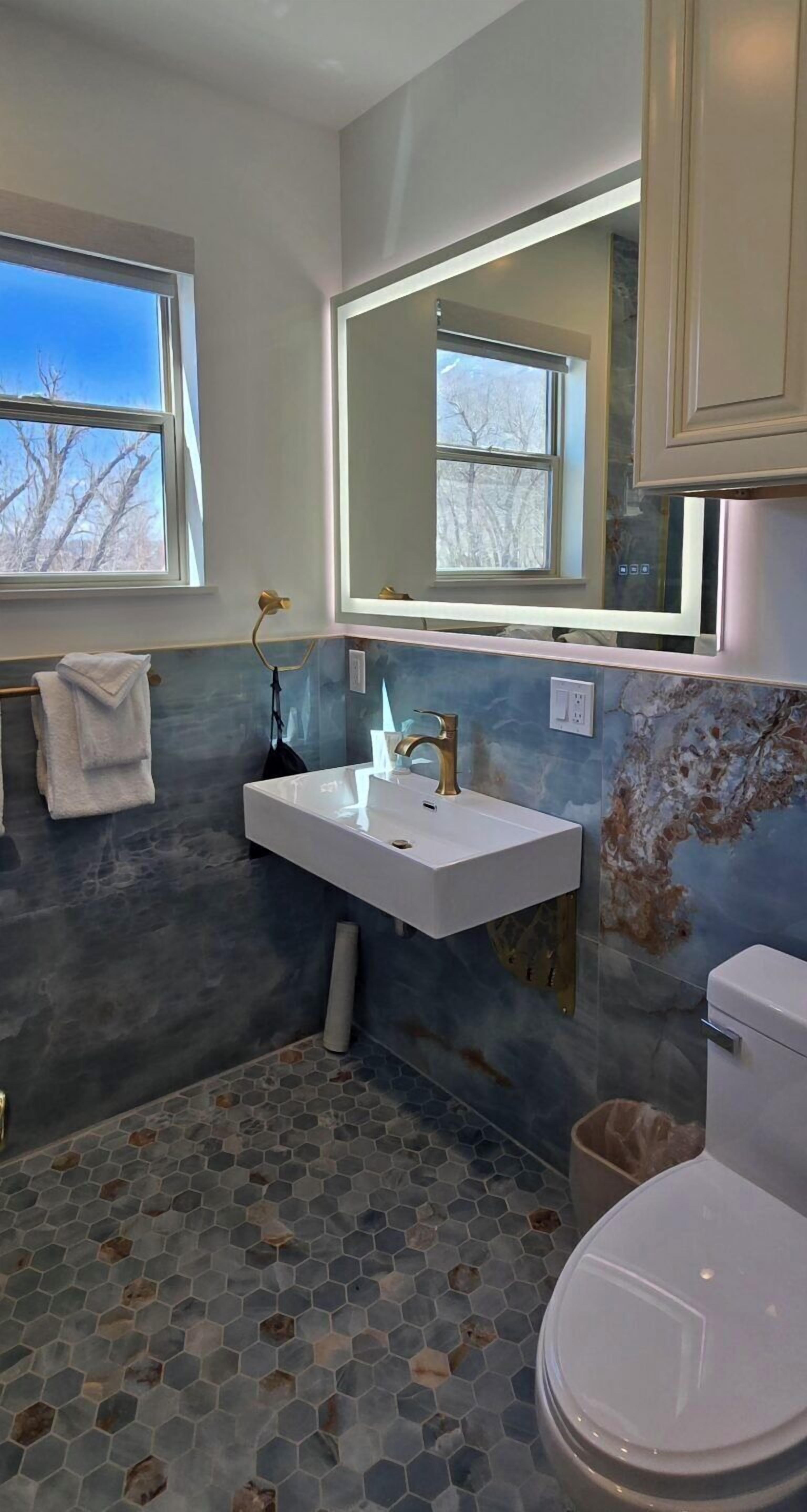 Un baño con un lavamanos blanco, un espejo y un inodoro. Hay una ventana con vista a árboles.