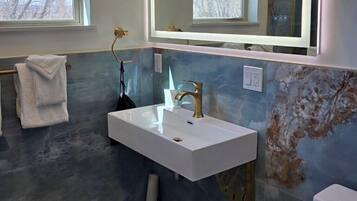 Un baño con un lavamanos blanco, un espejo y un inodoro. Hay una ventana con vista a árboles.