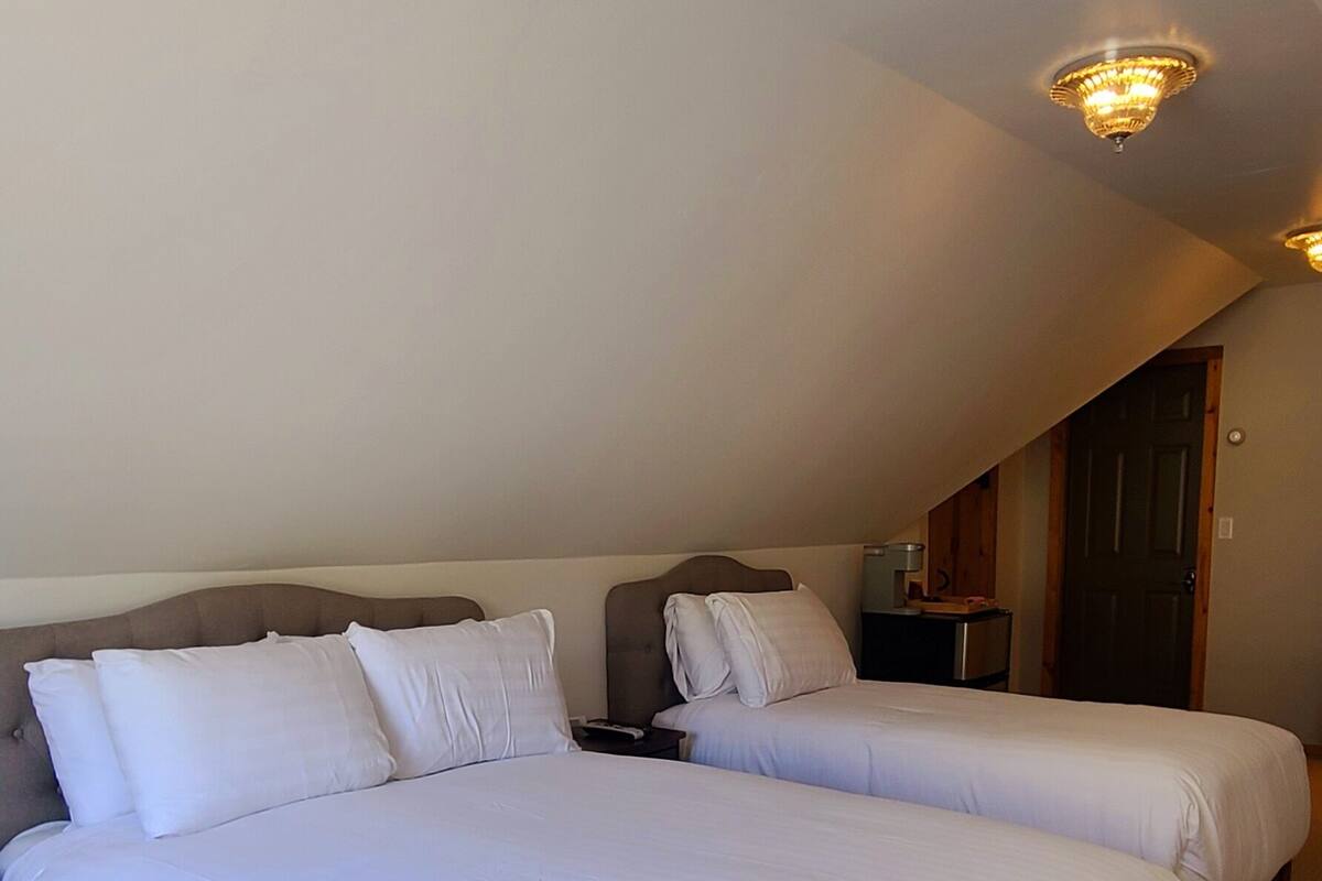 Quarto de hotel com duas camas, uma cabeceira de madeira, uma mesinha de cabeceira e um lustre no teto.