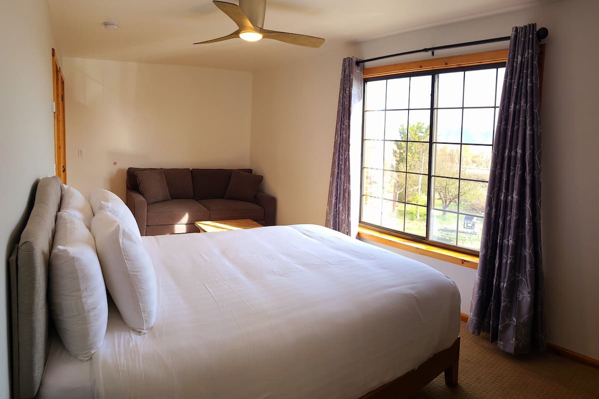 Um quarto com cama, ventilador de teto, janela com cortinas e um sofá.