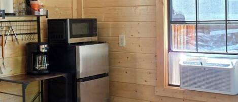 A compact room with a bed, a kitchenette with a refrigerator, a microwave, and a coffee maker. There's a window with blinds and a ceiling fan.
