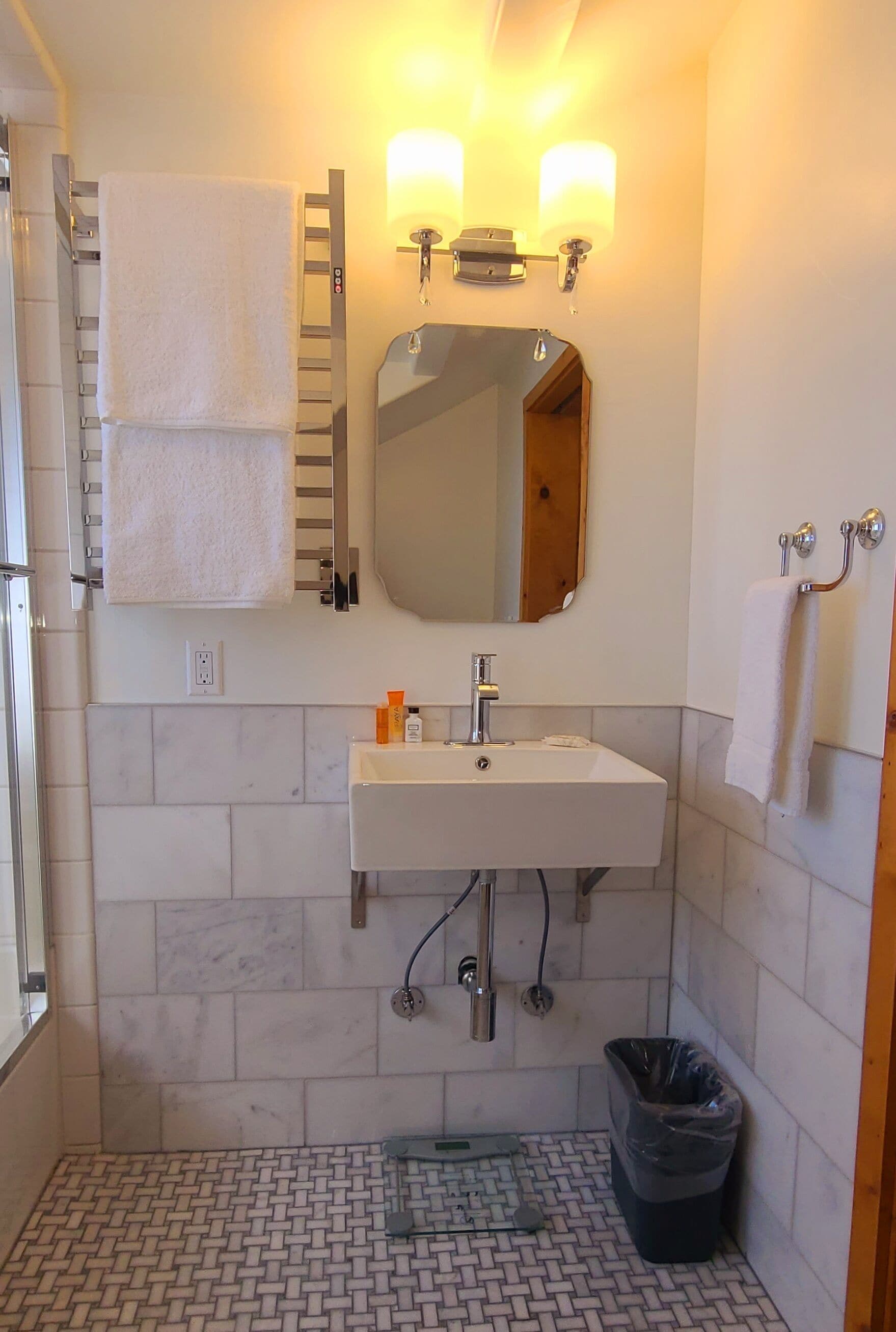 A bathroom with a white sink, mirror, and towel rack. There is a trash bin and a shower area.