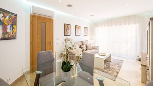Ein modernes Wohnzimmer mit einem Glastisch, grauen Sofas und einem beigen Sofa. Eine Holztür, eine weiße Klimaanlage und ein großes Fenster mit durchsichtigen Vorhängen sind ebenfalls zu sehen.