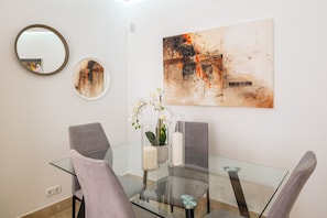 A dining area with a glass table, four chairs, a vase with flowers, and a large abstract painting on the wall.