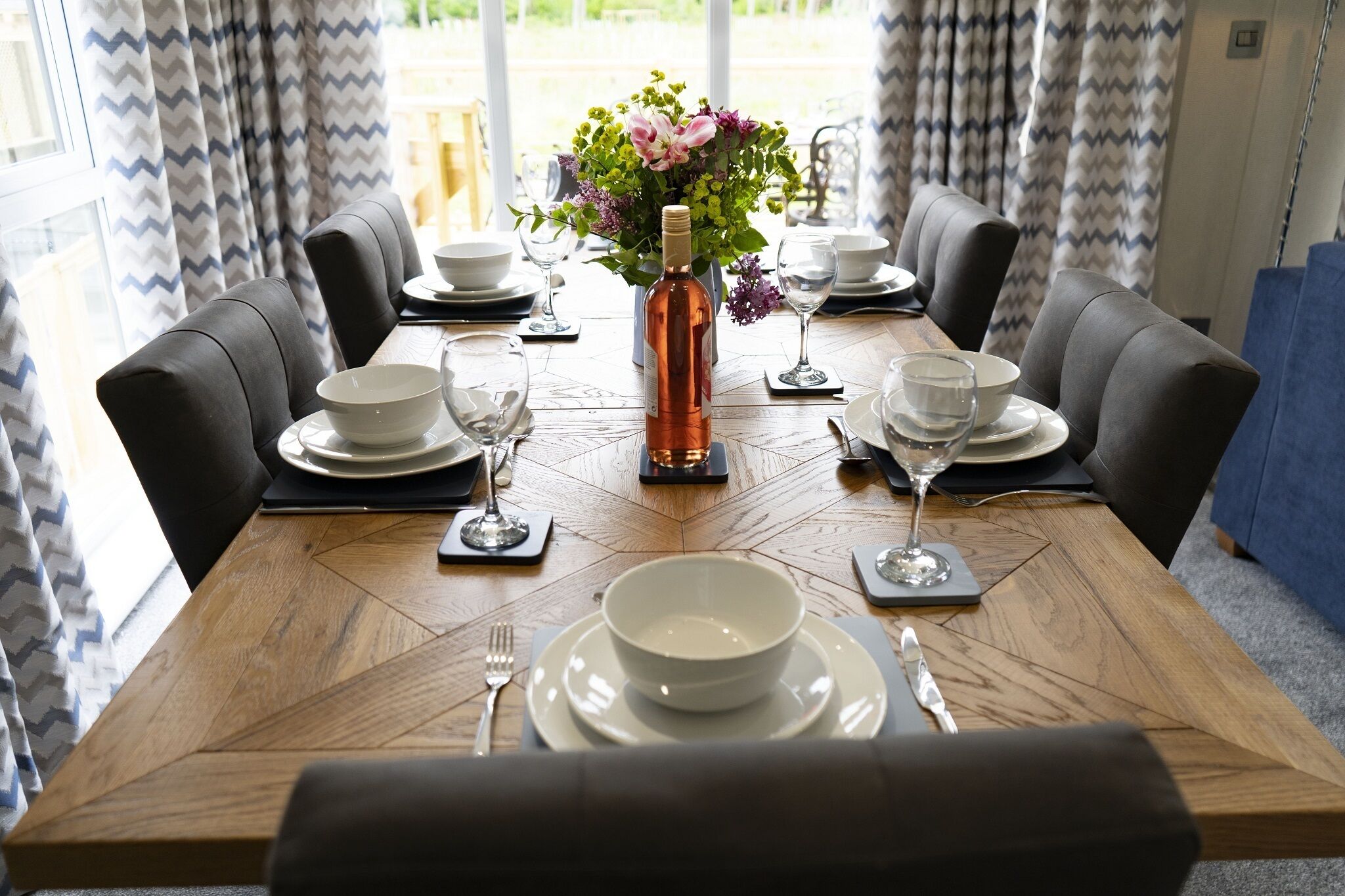 Mesa de comedor lista para seis personas, con un jarrón de flores, platos, tazones y copas de vino.