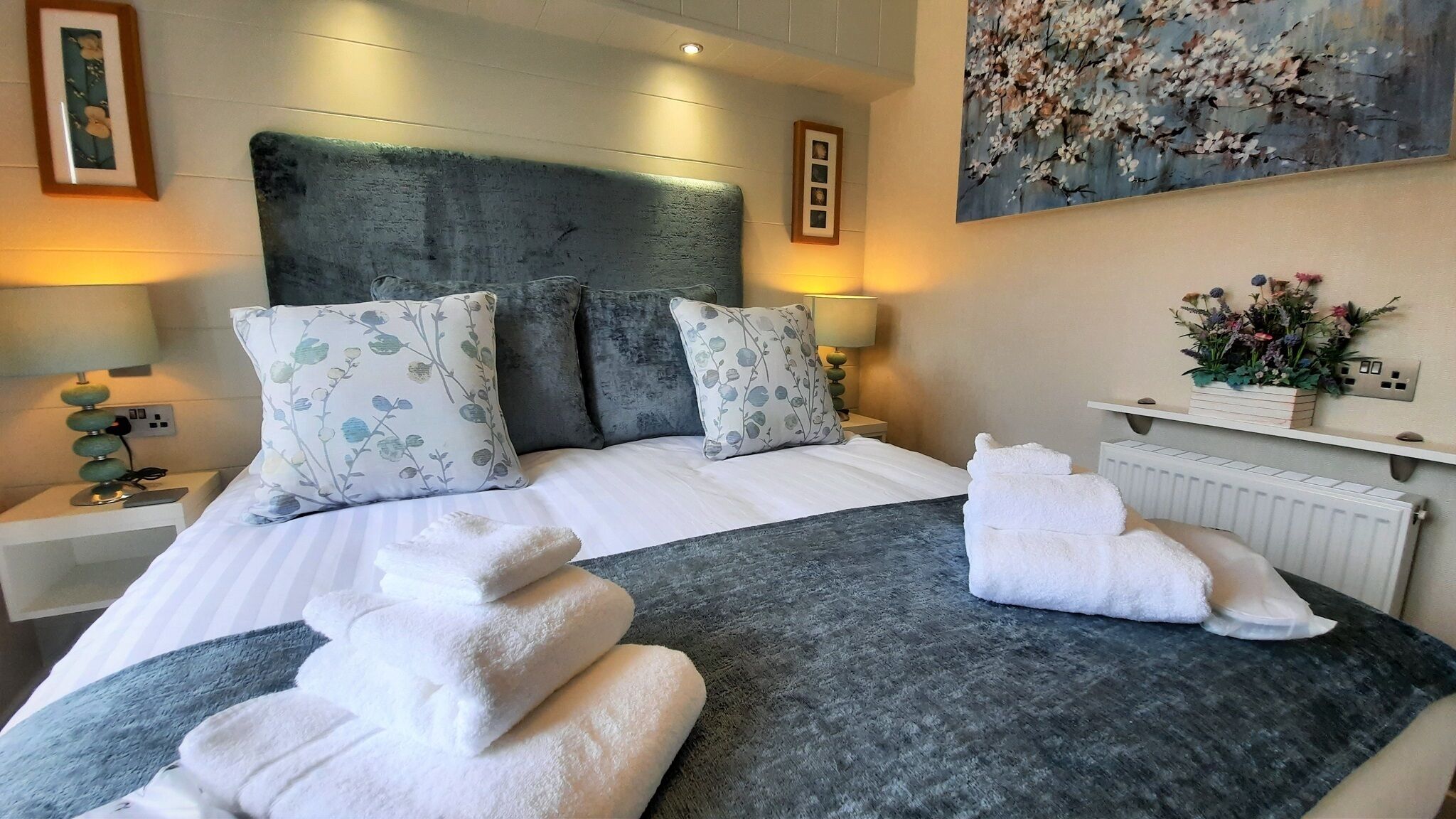 Una habitación de hotel con una cama grande, dos almohadas, una mesita de noche con una lámpara, un cuadro en la pared y un arreglo floral.