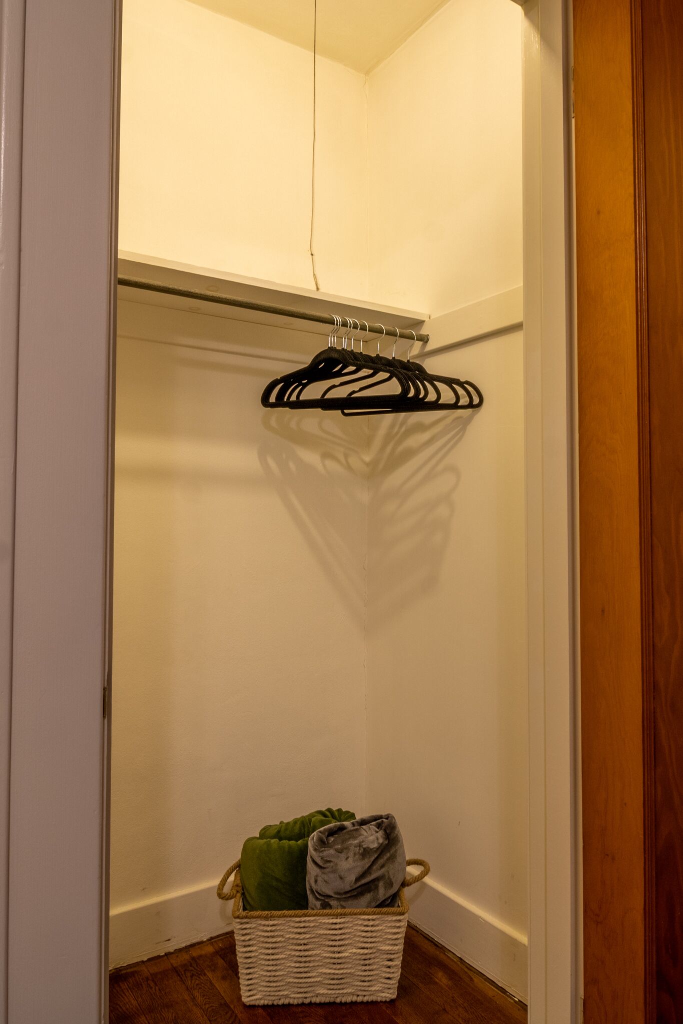 Een smalle gang met een houten deur aan de rechterkant en een witte muur aan de linkerkant. Een zwarte hanger hangt aan de muur, en er staat een rieten mand met een groene doek op de vloer.