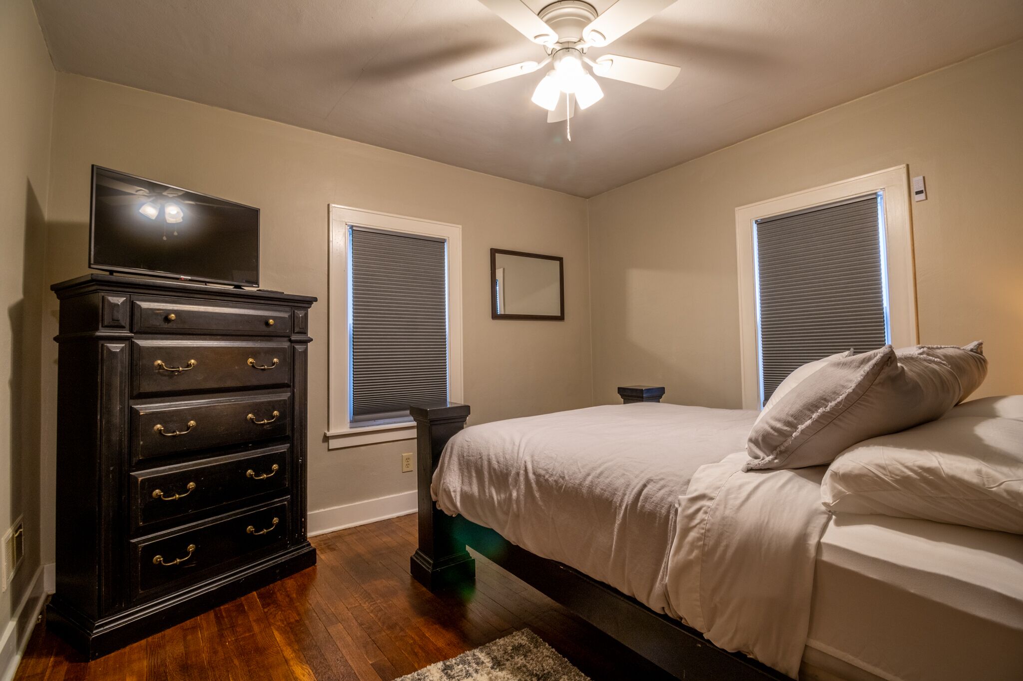 A bedroom with a bed, a dresser, a ceiling fan, and two windows with blinds.