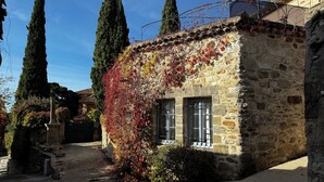 Un bâtiment en pierre avec une petite fenêtre et une porte, entouré d’arbres et bordé d’un chemin de gravier.