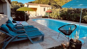 Un espace aménagé au bord de la piscine, avec des chaises longues, un pot de fleurs et une maison en arrière-plan.