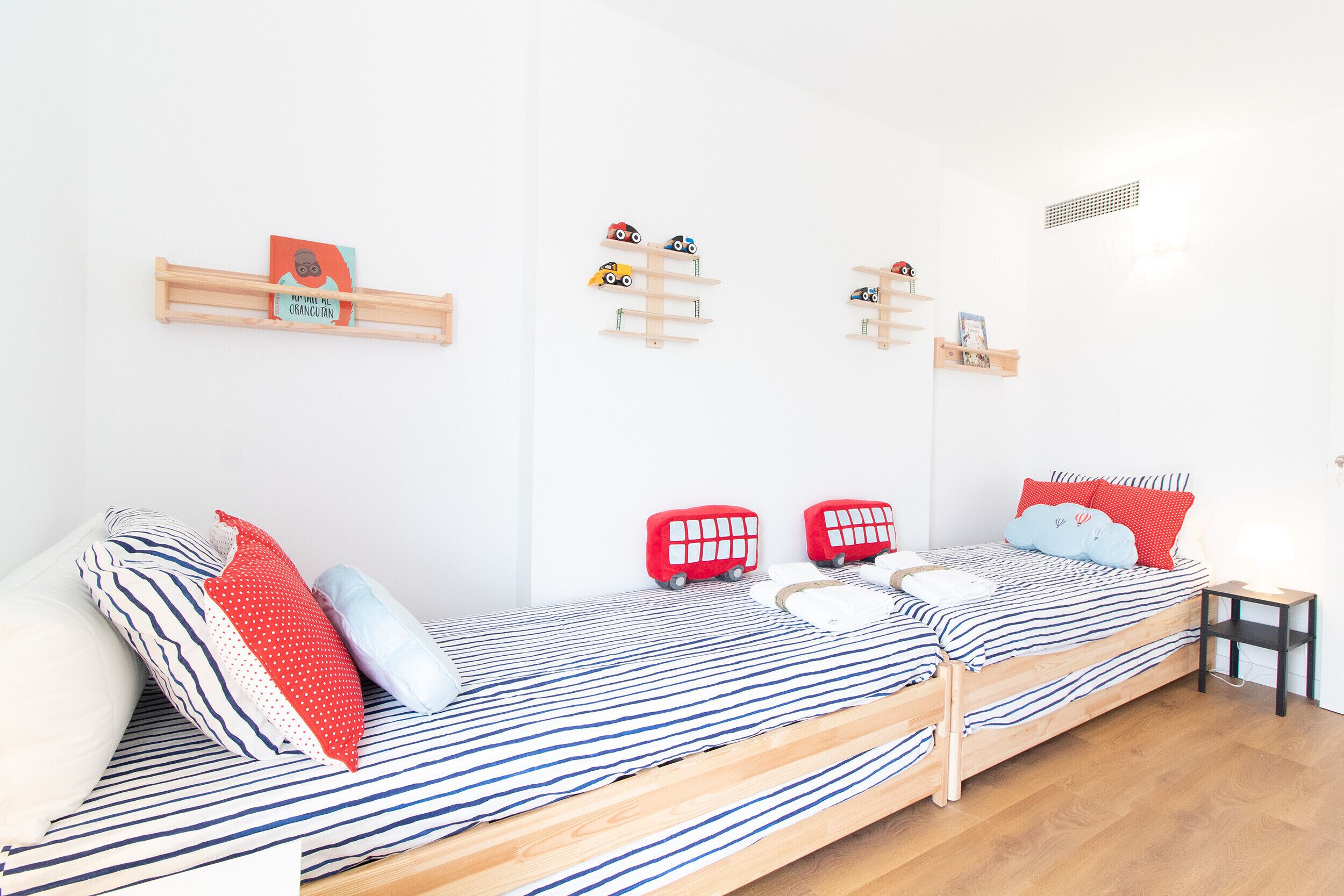 Une chambre avec un lit simple, une couvre-lit à rayures, deux oreillers, une petite table de chevet et une étagère fixée au mur, sur laquelle sont posés des jouets pour enfants.