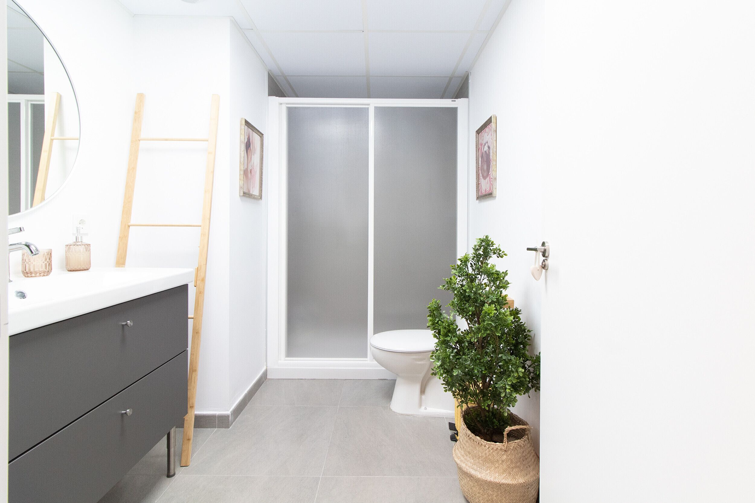 Un bagno con un mobile lavabo bianco, uno specchio tondo, una scala in legno, un water e una pianta in vaso.