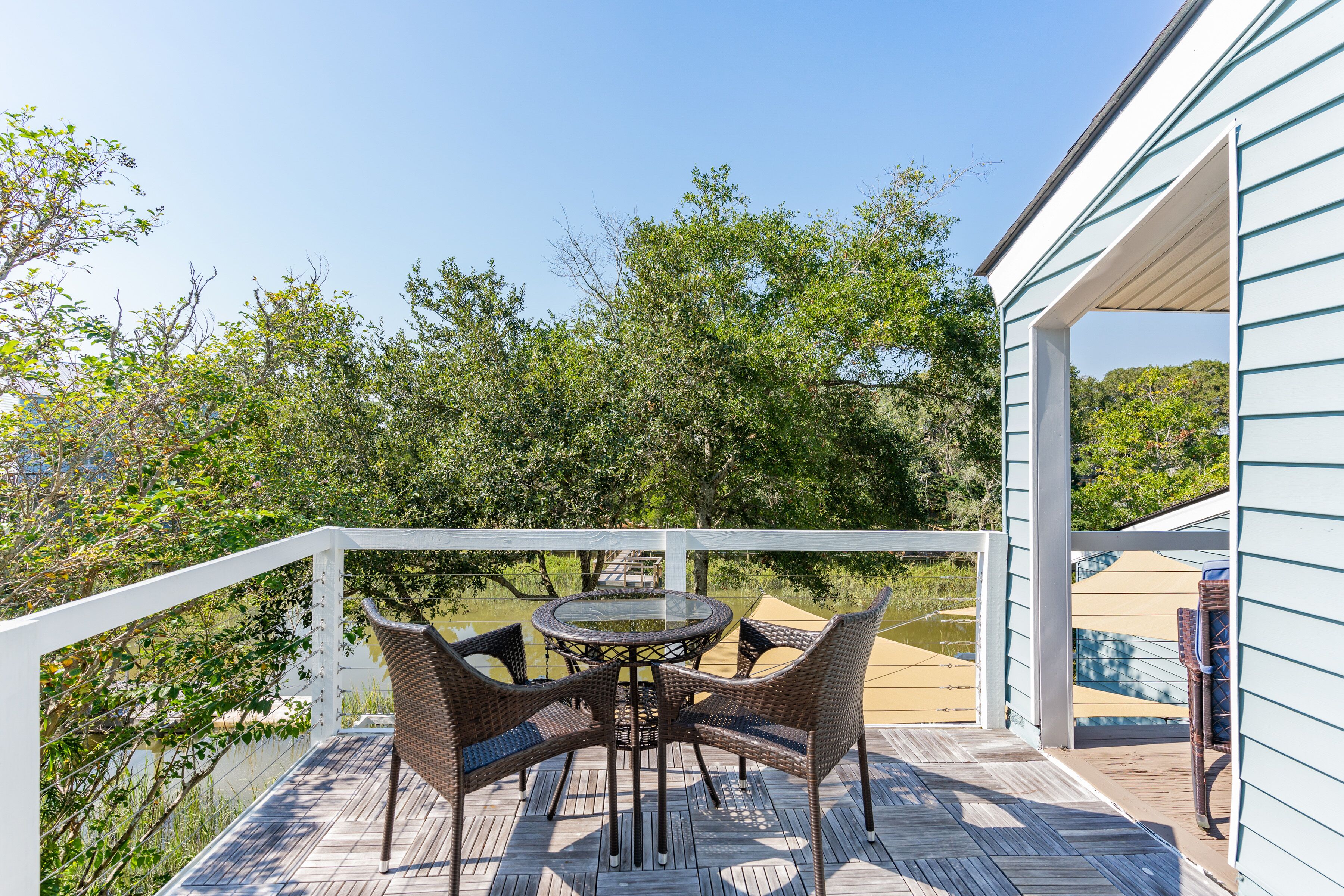 A wooden deck with a glass-top table and wicker chairs, overlooking a body of water and trees.