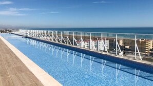 A long swimming pool with a glass railing, a clear blue sky, and a view of buildings and the sea in the background.