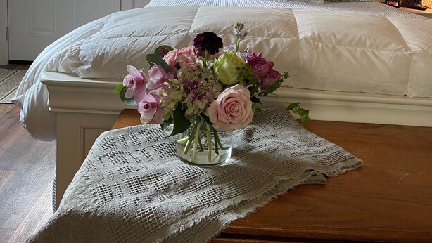 A bedroom with a bed, a bedside table with a vase of flowers, a mirror, and a door with a window.
