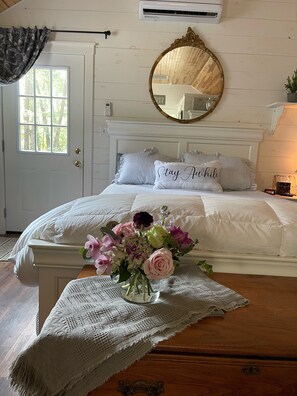 Une chambre à coucher avec un lit, une table de chevet ornée d’un vase de fleurs, un miroir et une porte avec une fenêtre.