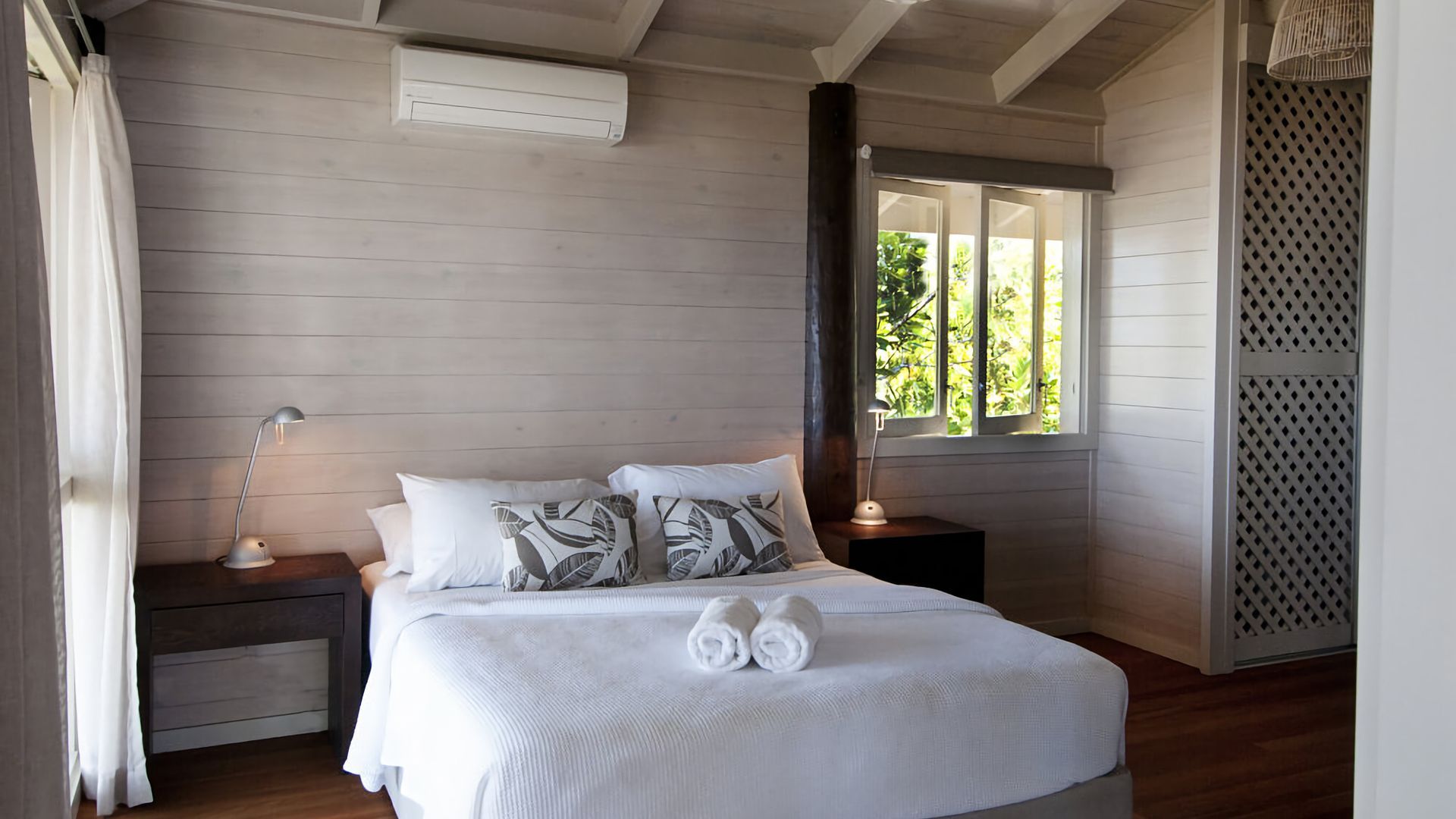 A bedroom with a bed, two bedside tables, a window, and a wall-mounted air conditioner.