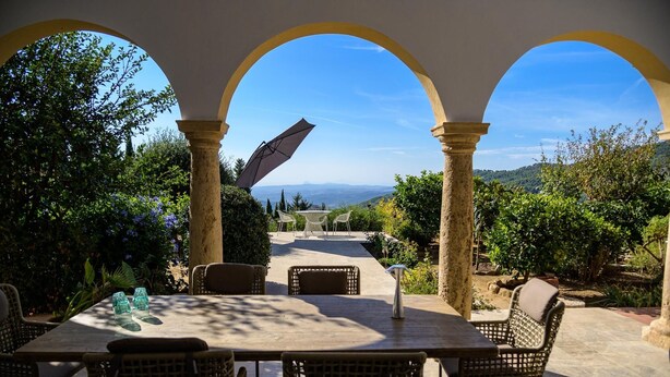 Eine Terrasse im Freien mit Tisch und Stühlen, umgeben von Bögen und Grünpflanzen, mit Blick auf eine schöne Landschaft.