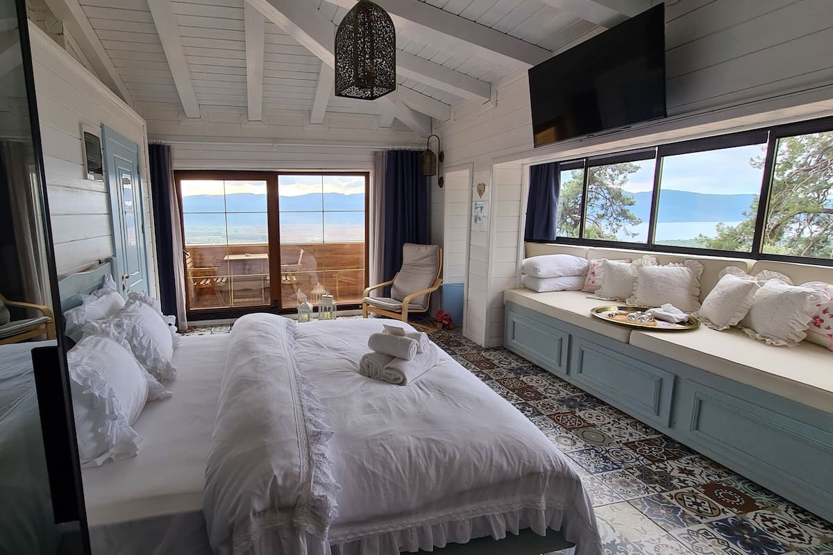 Un dormitorio con una cama grande, un asiento junto a la ventana con almohadas, una silla y vistas al exterior.