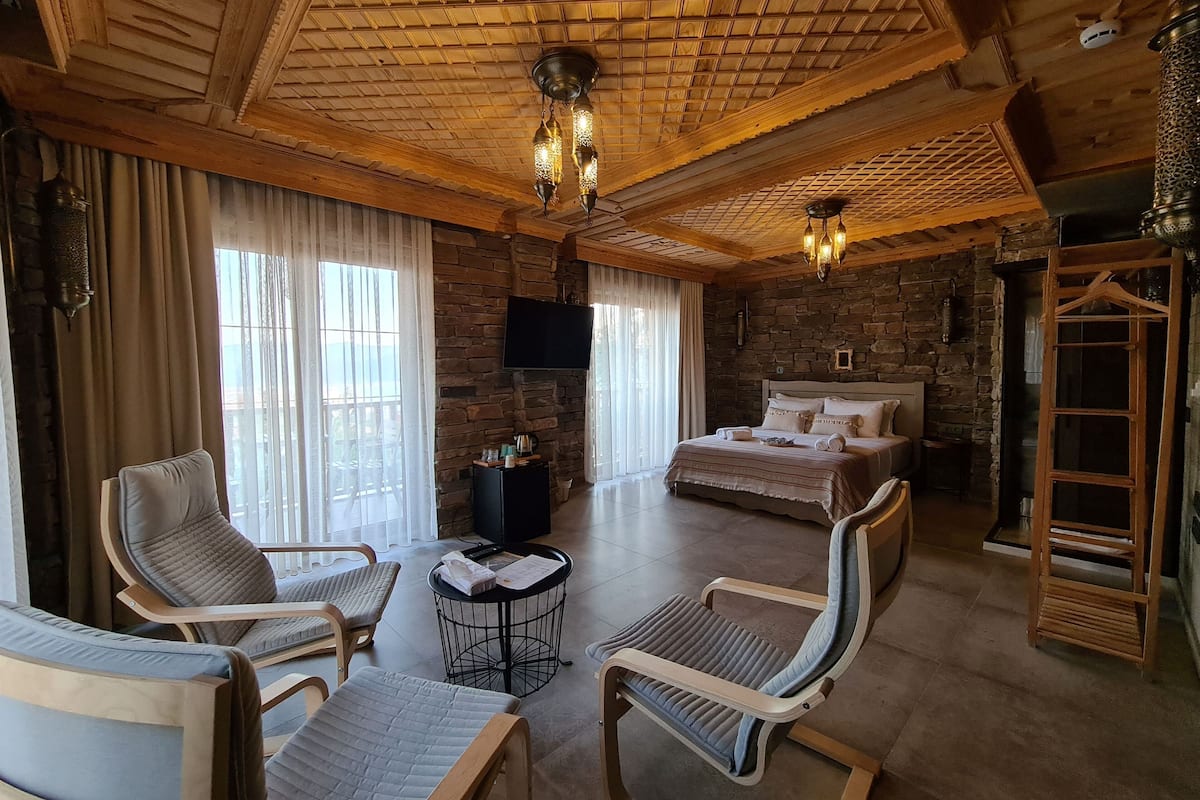 Una habitación amplia con una cama grande, dos sillones, una mesita y un televisor. La habitación cuenta con vigas de madera en el techo, paredes de piedra y un ventanal con cortinas.