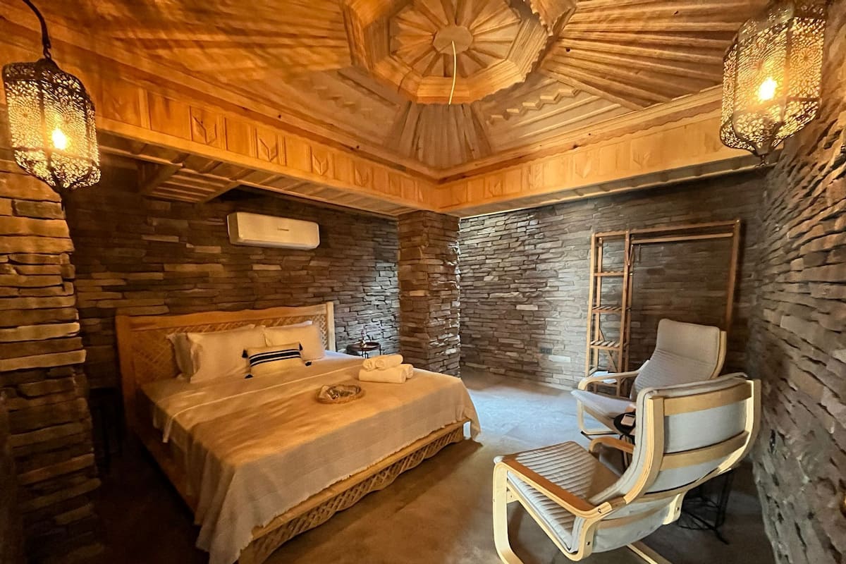 Un dormitorio con pared de piedra, cama, sillón y ventilador de techo.