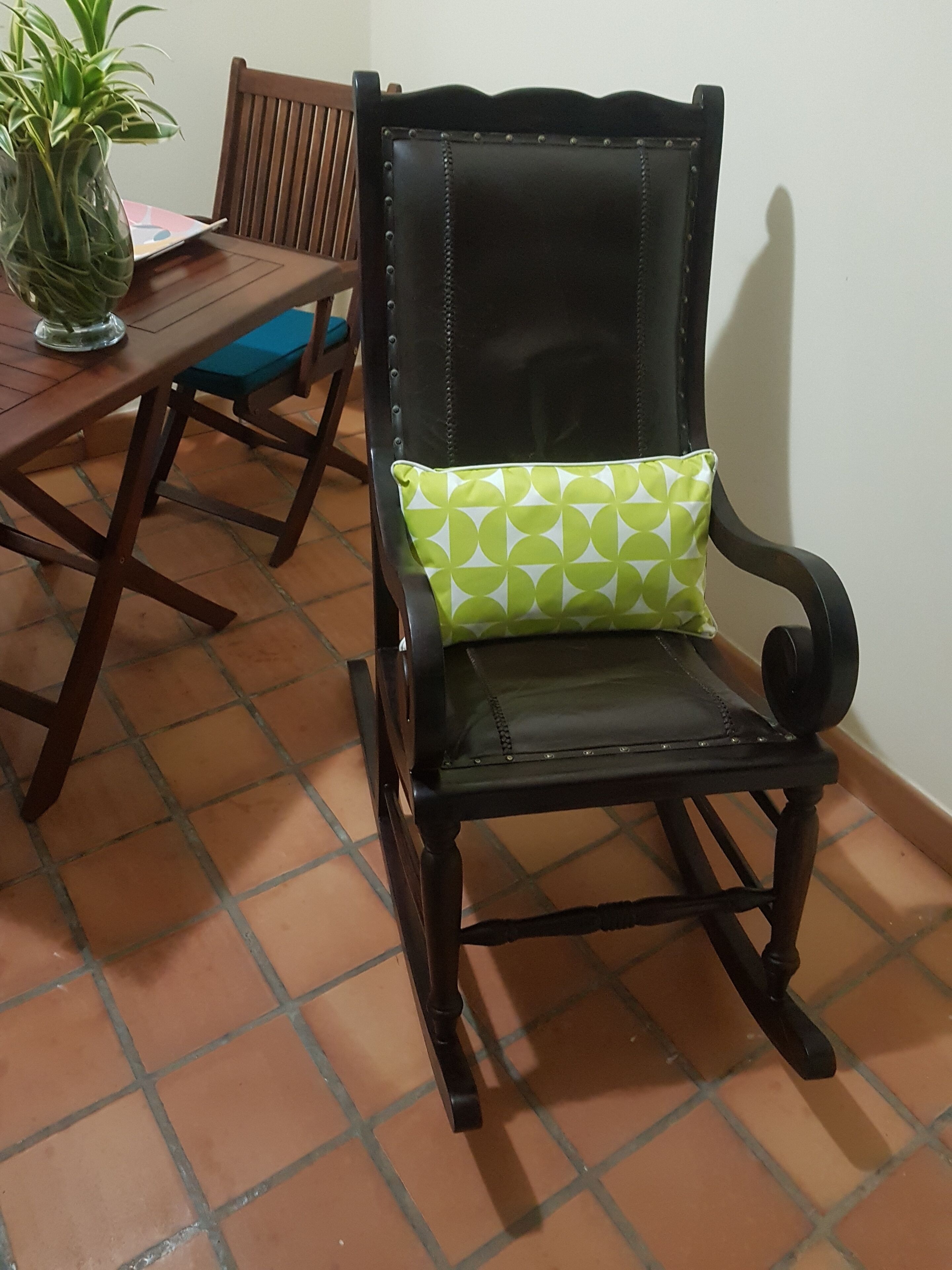 A chair with a green patterned cushion, a wooden table with a vase of green plants, and a tiled floor.