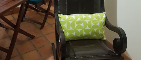 A chair with a green patterned cushion, a wooden table with a vase of green plants, and a tiled floor.