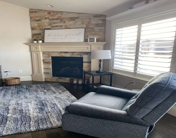 A living room with a stone fireplace, a gray armchair, a blue rug, and a window with blinds. - Cheerful, spacious, sunny & comfy 4-bdrm home (Rock Springs)