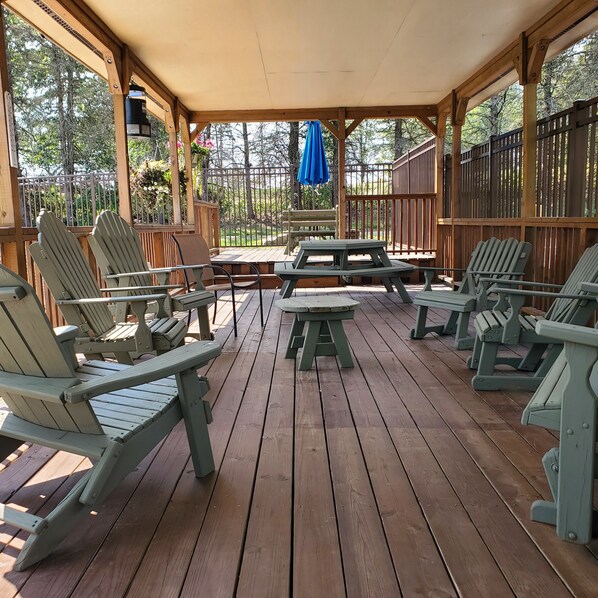 A covered outdoor seating area with wooden chairs and a picnic table, surrounded by a wooden fence and trees.