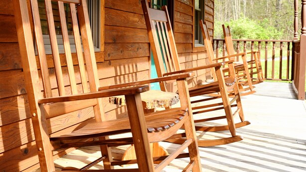 Een houten schommelstoel op een veranda, met een houten hutje op de achtergrond.