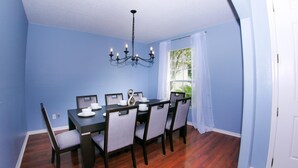 A dining room with a dark wooden table and matching chairs, a chandelier, and a window with white curtains.