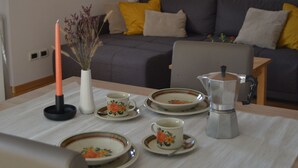 Un comedor con una mesa dispuesta para dos personas, que incluye platos, una taza, una vela y un florero con flores secas. Un sofá gris con cojines amarillos se ve al fondo.