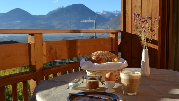 Una mesa de madera con una cesta de pan, una taza de café, un tarro de mermelada y un florero con flores en un balcón con vistas a montañas.