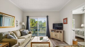 A living room with a sofa, coffee table, TV, and a balcony with a view of trees. - Great cozy beach getaway (Santa Rosa Beach)
